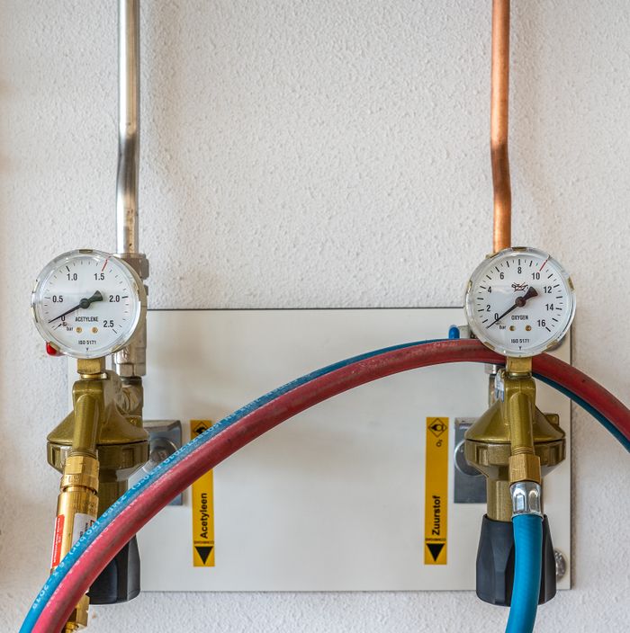 hero-img-01 Close-up of industrial gas pressure gauges connected with hoses against a white wall.